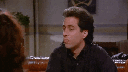 a man in a black shirt is sitting on a couch talking to a woman in a living room .