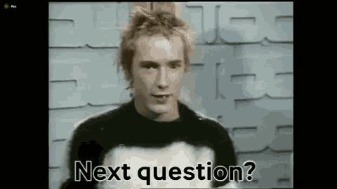 a man in a black and white sweater is asking a question while standing in front of a brick wall .