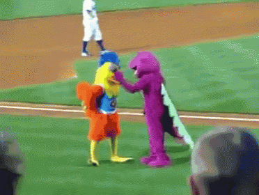 a baseball player stands on the mound while a mascot holds a purple blanket
