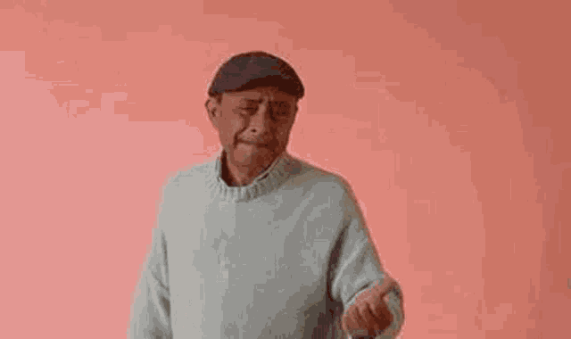 an elderly man wearing a hat and sweater is standing in front of a pink wall .