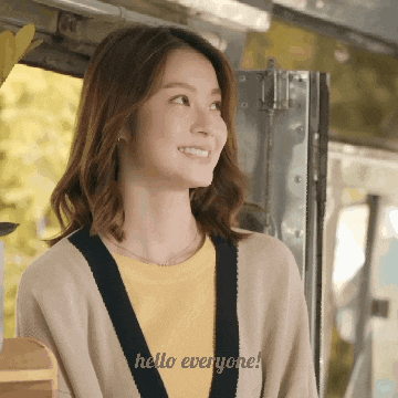 a woman wearing a yellow shirt and a tan cardigan is smiling and says hello everyone