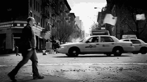 a black and white photo of a man walking down a city street next to a taxi .