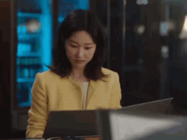 a woman in a yellow jacket is sitting at a table with a laptop and looking at the camera .