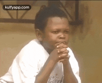 a young boy in a white shirt is sitting with his hands folded in front of his face .