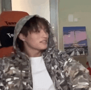 a young man wearing a camouflage jacket and a hoodie is sitting in a chair .