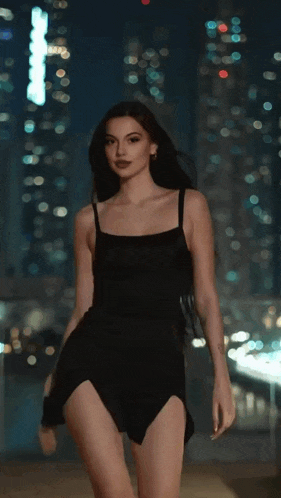 a woman in a black dress is standing in front of a city skyline at night