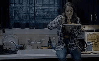 a woman in a plaid shirt is sitting on a kitchen counter eating a plate of food