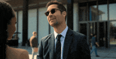 a man in a suit and tie wearing sunglasses is talking to a woman