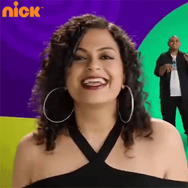 a woman wearing hoop earrings and a black top is smiling in front of a nick logo
