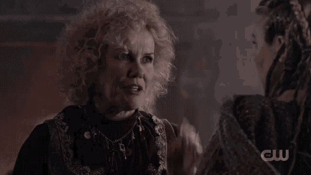 a woman with curly hair is talking to another woman with a cw logo behind her
