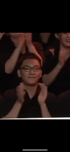a man wearing glasses is sitting in a crowd of people applauding .