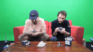 two men are sitting on a red couch playing a game with a green screen behind them