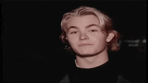 a young man with blonde hair and a black shirt is making a funny face in a dark room .