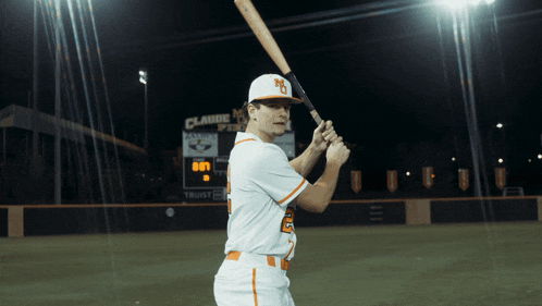 a baseball player is holding a bat in front of a scoreboard that says claude field