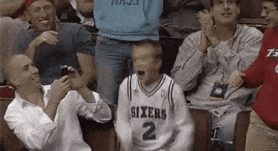 a man in a sixers jersey is sitting in a crowd of people watching a basketball game .