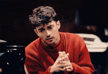 a man in a red sweater sits with his hands folded in front of a piano