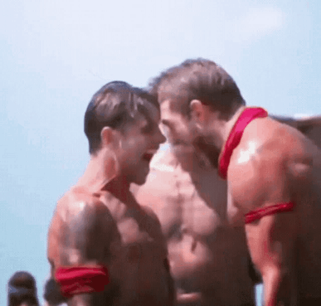 two shirtless men are standing next to each other on a beach and talking to each other .
