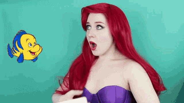 a woman with red hair is dressed as ariel from the little mermaid and looking at a fish .