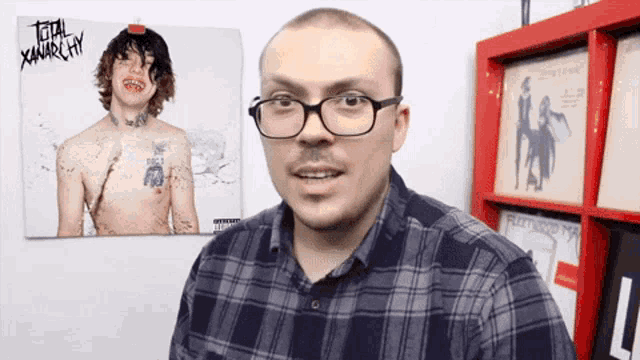 a man wearing glasses stands in front of a picture of a shirtless man with the words total anarchy on it