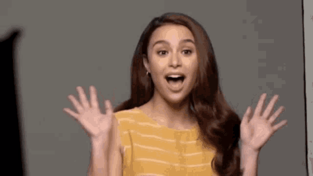 a woman in a yellow striped shirt is making a surprised face with her hands up .