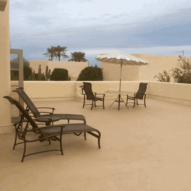 a patio with chairs a table and an umbrella with palm trees in the background