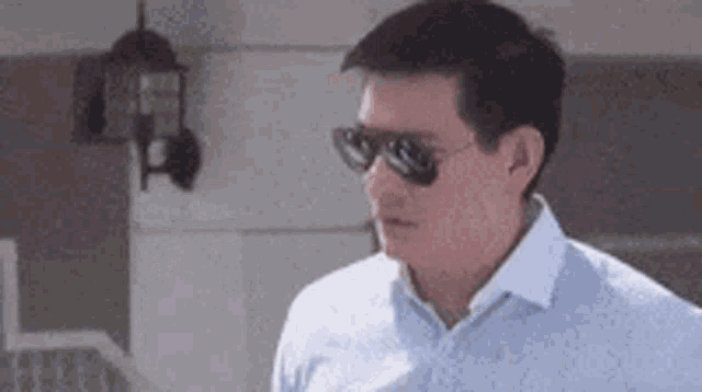 a man wearing sunglasses and a blue shirt is standing in front of a house .