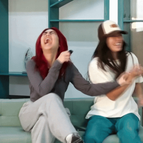 two women are sitting on a couch laughing with one wearing a hat