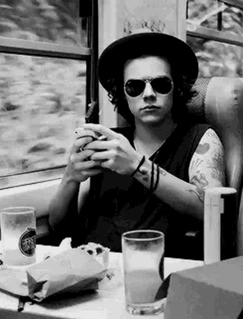 harry styles is sitting on a train wearing a hat and sunglasses .