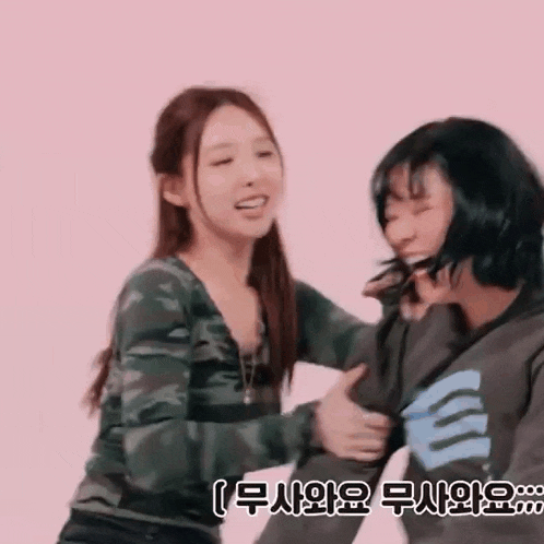 two women are hugging each other on a pink background . one of the women is wearing a camouflage shirt .
