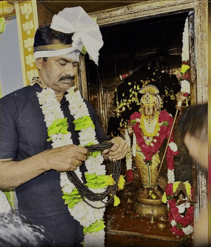 a man wearing a turban holds a necklace in front of a statue of a deity