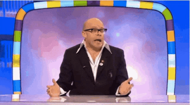 a bald man in a suit and glasses is sitting at a table with his hands outstretched .