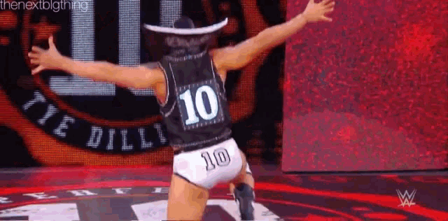 a wrestler wearing a cowboy hat and a jersey with the number 10 on it is kneeling on a stage .