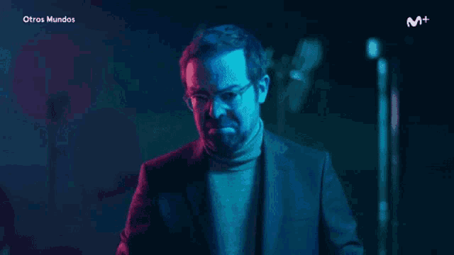 a man in a suit and turtleneck stands in front of a blue and red background that says otros mundos