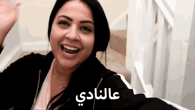 a woman in a black jacket is smiling in front of stairs with arabic writing on the bottom