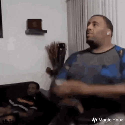 a man is sitting on a couch in a living room while another man sits on a couch in the background .