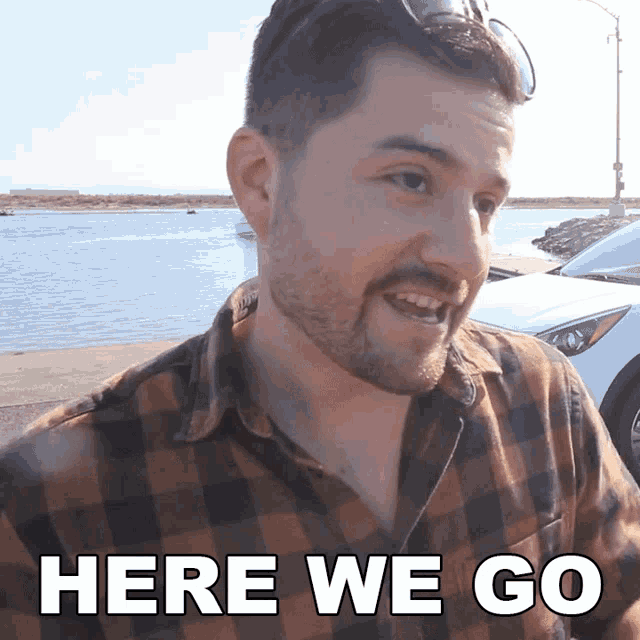 a man in a plaid shirt says " here we go " in front of a body of water