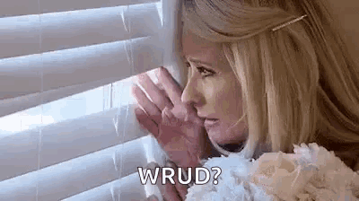 a woman is looking out of a window with blinds and holding her hand to her face .
