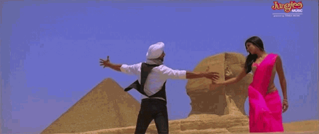 a man in a white turban and a woman in a pink saree are dancing in front of pyramids sponsored by junglee