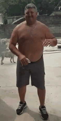 a shirtless man in shorts and sneakers is standing in a driveway with a dog in the background .