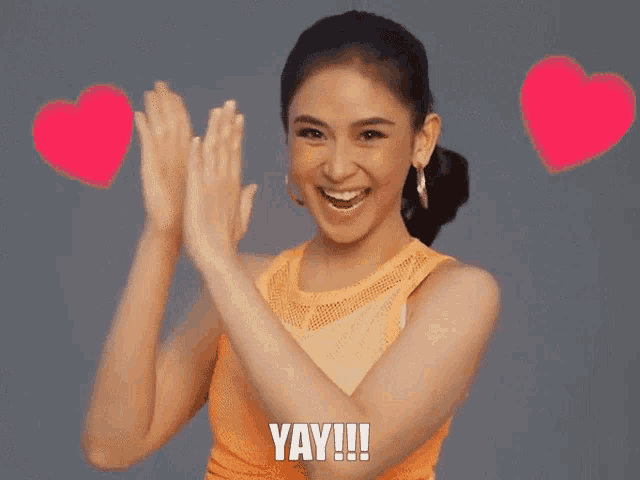 a woman in an orange tank top is clapping her hands and saying yay