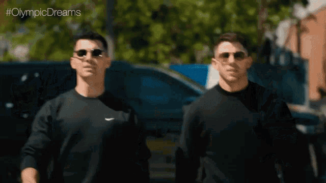two men wearing black sweaters and sunglasses are walking down a street .