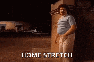 a man with a beard is standing in front of a building with the words `` home stretch '' written on the screen .