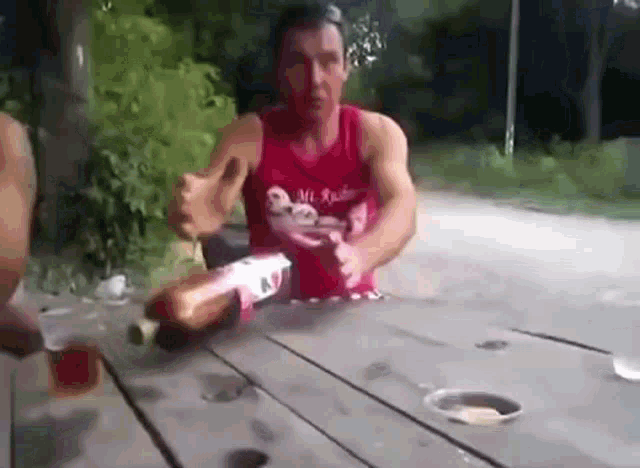 a man in a red tank top is sitting at a picnic table holding a bottle of alcohol .