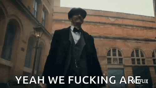 a man in a suit and bow tie is standing in front of a building and says yeah we fucking are .