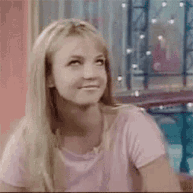 a blonde woman wearing a pink shirt is smiling and looking at the camera .