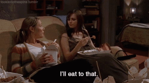 two women are sitting on a couch eating food and one of them is saying i 'll eat to that