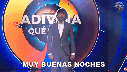a man in a suit stands on a stage in front of a sign that says adivina que muy buenas noches