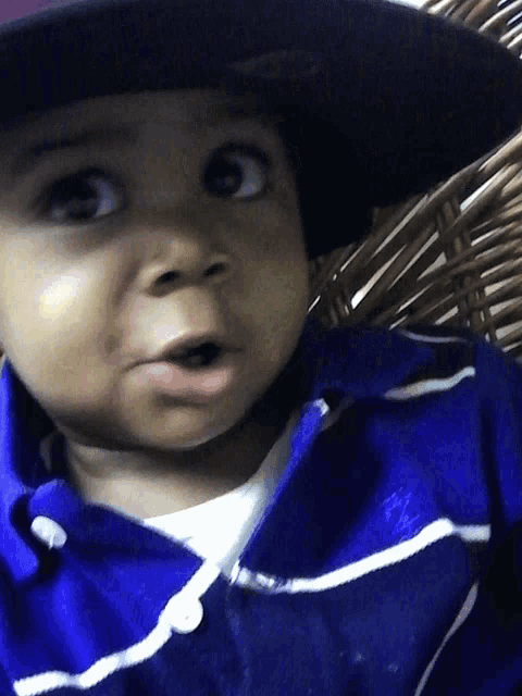 a baby wearing a black hat and a blue shirt is sitting in a wicker chair