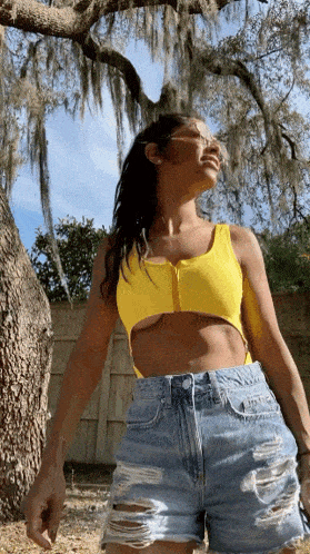a woman in a yellow crop top and denim shorts is standing under a tree .
