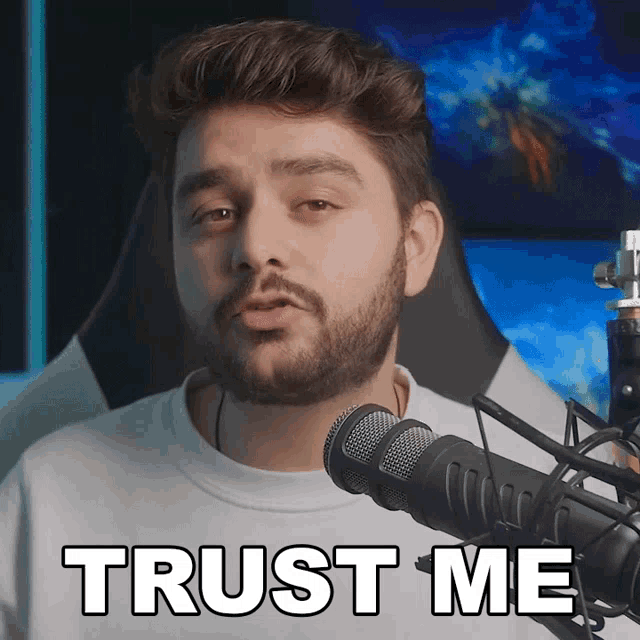 a man with a beard stands in front of a microphone with the words trust me above him